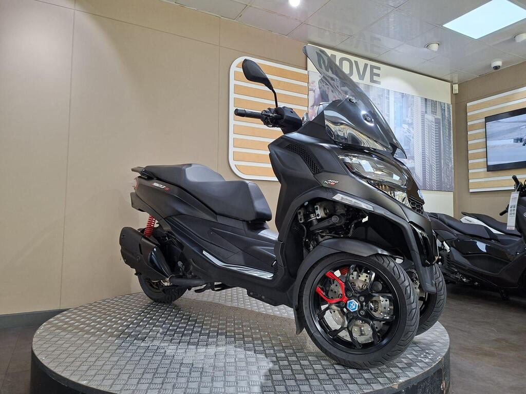 Piaggio MP3 400 LC72 KHB 20 A sleek black scooter with three wheels is displayed on a circular platform inside a showroom. The scooter features modern design elements, including a windshield and prominent red brake calipers. Background displays include wall panels and an additional scooter.