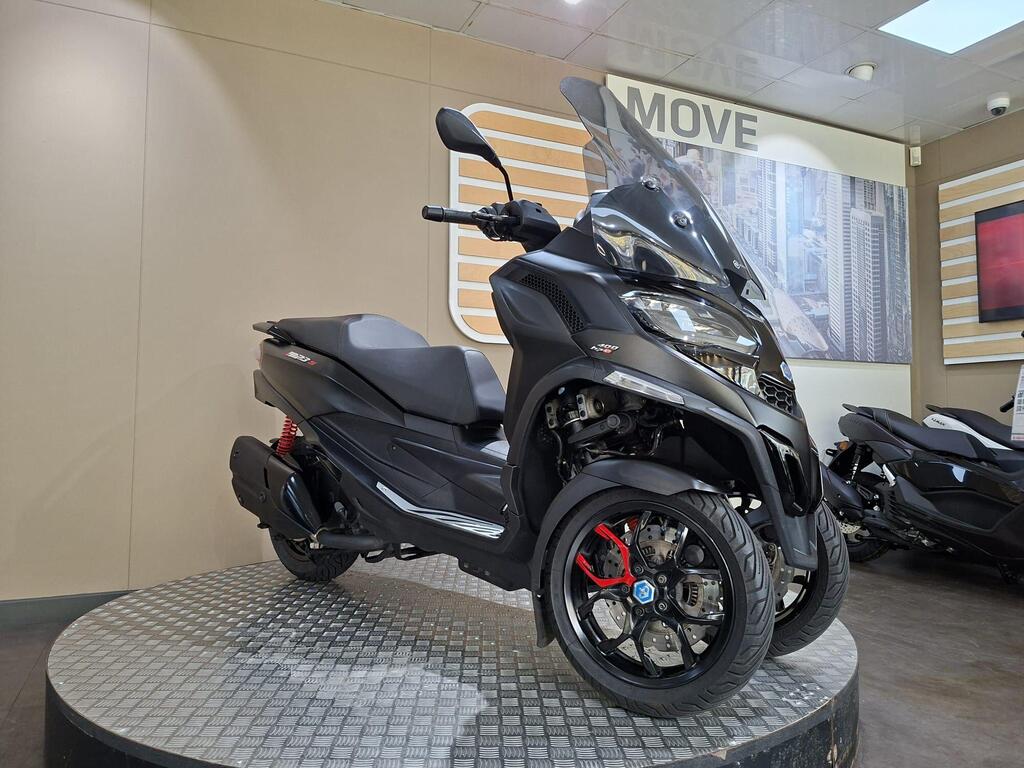 Piaggio MP3 400 LC72 KHB 21 A black three-wheeled scooter with a transparent windscreen displayed on a round platform in a showroom, featuring a modern design and red accents on the wheels. The background includes a wall with a cityscape image and the word "MOVE."