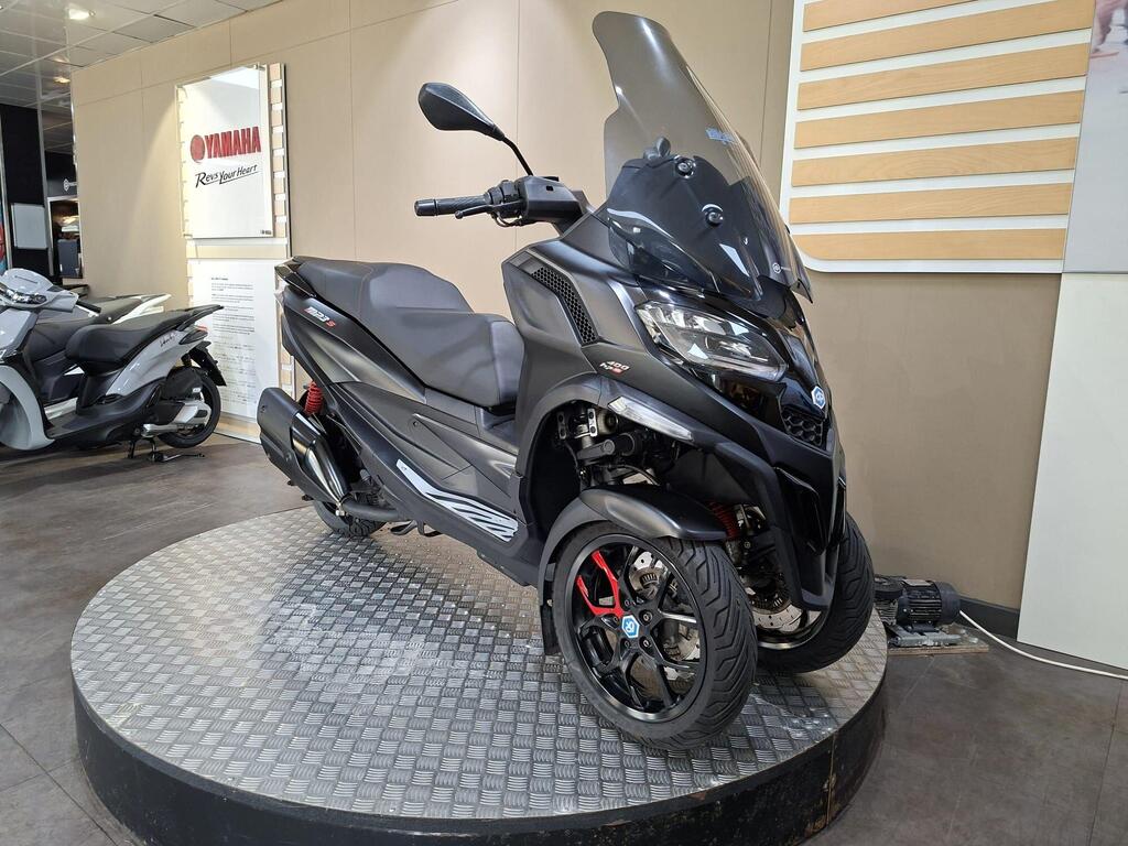 Piaggio MP3 400 LC72 KHB 3 A black three-wheeled scooter displayed on a circular platform in a Yamaha showroom, featuring a large windshield and sporty design elements. A second scooter is partially visible in the background.