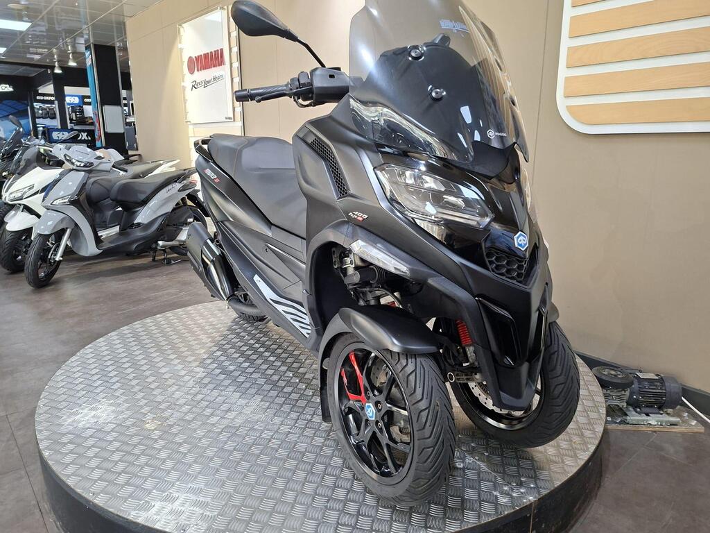 Piaggio MP3 400 LC72 KHB 4 A sleek black three-wheeled scooter displayed on a circular showcase in a dealership, with other scooters visible in the background. The scooter features sporty design elements and red accents on the wheels.
