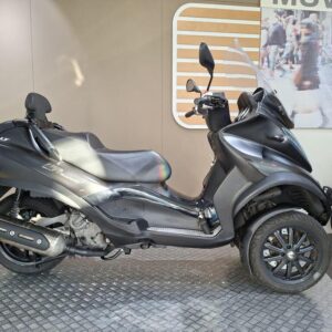 Piaggio MP3 500 Sport Touring LT (2017) A black three-wheeled scooter displayed on a textured circular platform, with a modern backdrop featuring a city scene. The scooter has a windshield and a sleek design, showcasing its sporty aesthetics.