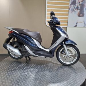 Piaggio Medley 125 2019 A blue and silver scooter parked on a circular platform, with a neutral-colored background and a blurred image of people walking in the distance.