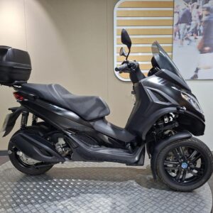 Piaggio MP3 300 HPE (2024) A sleek black three-wheeled scooter displayed on a textured platform, featuring a top box for storage and a windshield for protection. The background includes a blurred image of people walking.