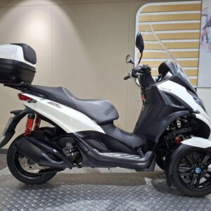 Piaggio MP3 300 HPE Sport (2022) A white and black three-wheeled scooter is parked on a circular metal platform in a showroom, featuring a top case and stand, with a neutral-colored wall and an abstract image of pedestrians in the background.