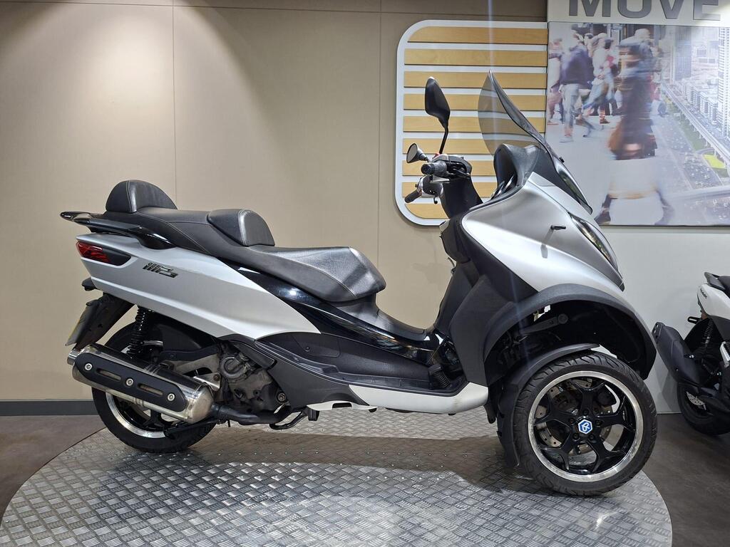 Piaggio MP3 500 2016 1 Piaggio MP3 500 LT Sport (2016) Side view of a silver scooter with dual front wheels, featuring a black seat and sporty design, displayed on a circular metal platform in a modern showroom setting.