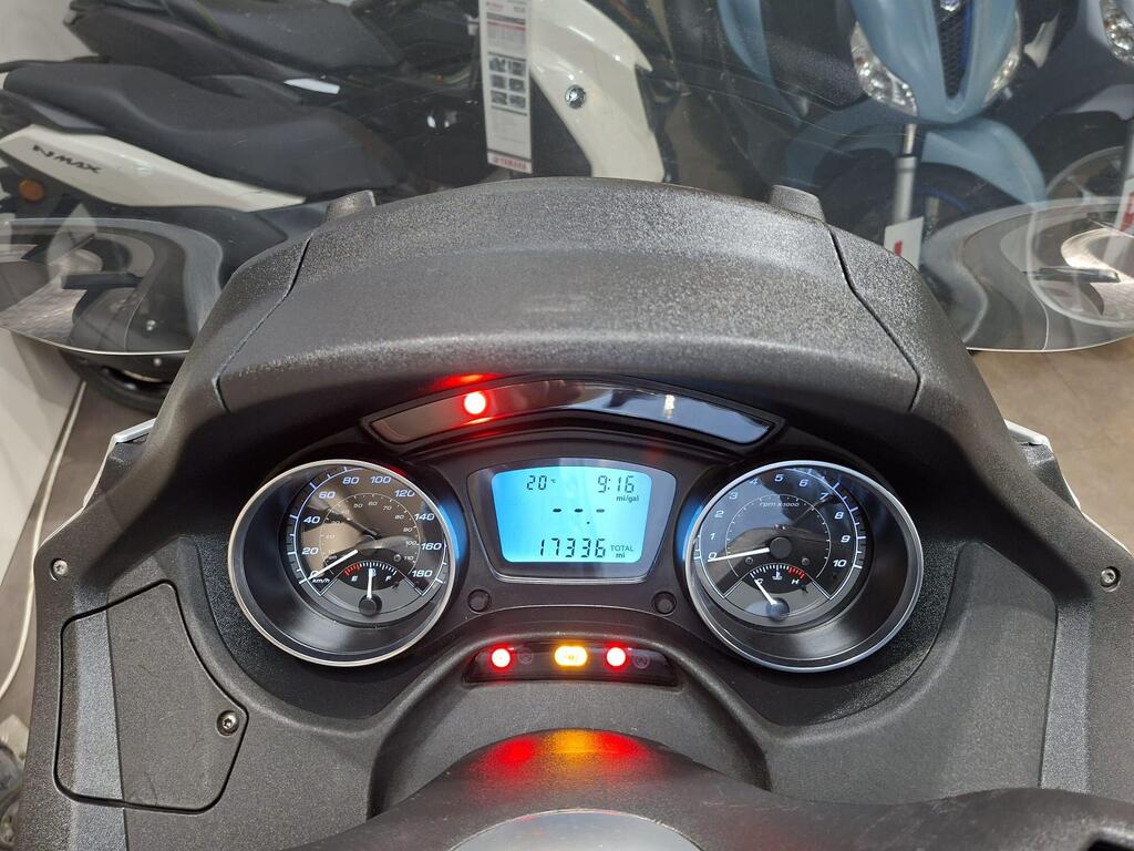 Piaggio MP3 500 2016 10 Close-up view of a scooter dashboard displaying speedometer and odometer readings, with illuminated indicator lights, and a digital screen showing time and mileage. The background features blurred scooters.