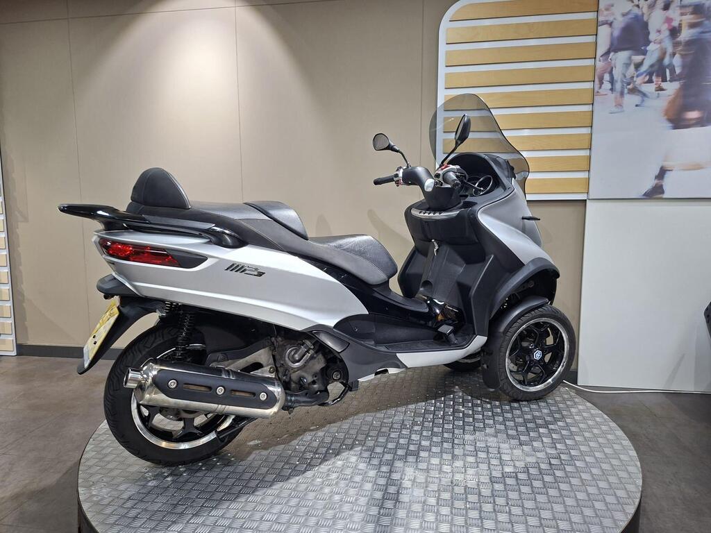 Piaggio MP3 500 2016 11 A silver and black three-wheeled scooter displayed at an exhibition, featuring a sleek design, large windshield, and a rounded rear. The scooter is positioned on a textured surface with a neutral background.