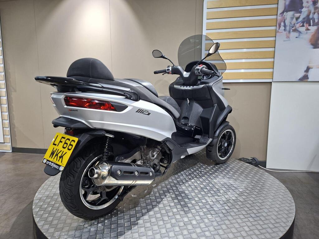 Piaggio MP3 500 2016 12 A white scooter displayed on a circular platform, showing its rear profile with a black seat and sleek design. The vehicle features a visible license plate reading "LF66 WXY." The background includes neutral-colored walls and a decorative panel.