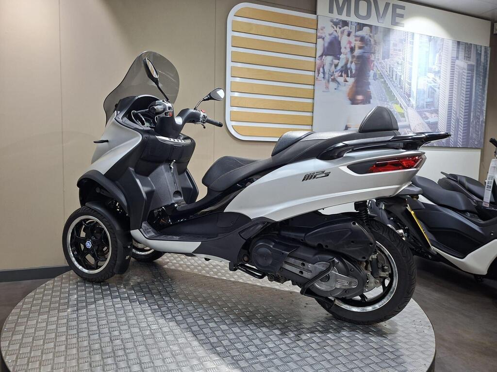 Piaggio MP3 500 2016 13 A sleek white and black three-wheeled scooter displayed on a rotating platform in a modern showroom, with a backdrop featuring cityscape imagery.