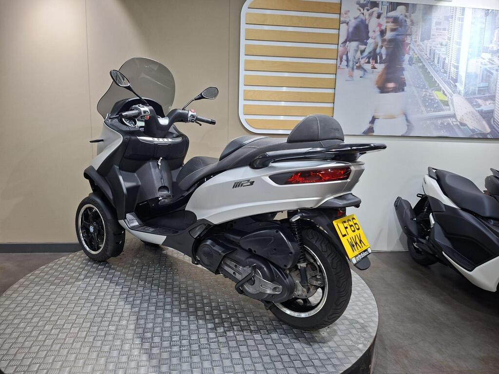 Piaggio MP3 500 2016 14 A white three-wheeled scooter displayed on a circular platform in a showroom, with a clear windshield and black seating. The background features a large image of a busy city street.