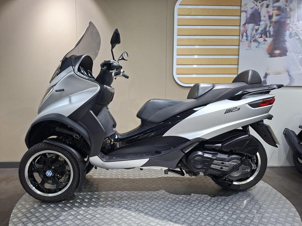 Piaggio MP3 500 2016 15 A side view of a silver three-wheeled scooter with a black seat, displayed on a metal platform against a neutral background.