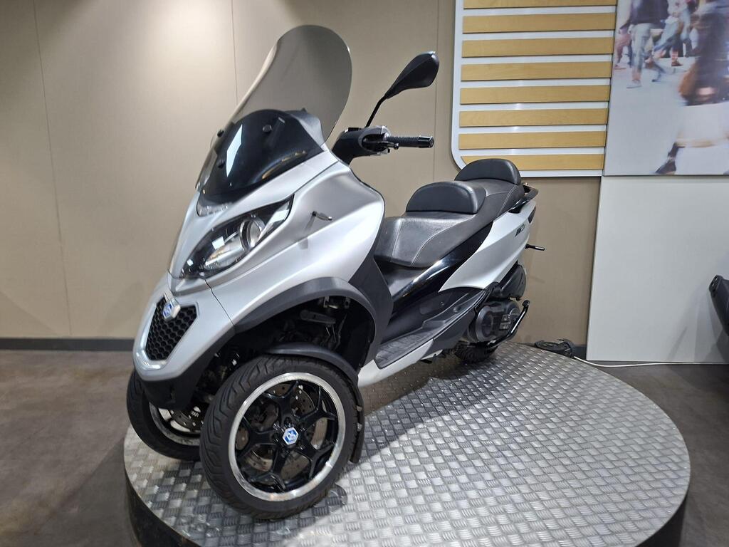 Piaggio MP3 500 2016 16 A silver three-wheeled scooter displayed on a circular, textured platform against a neutral background. It features a large windscreen and comfortable seating for two.