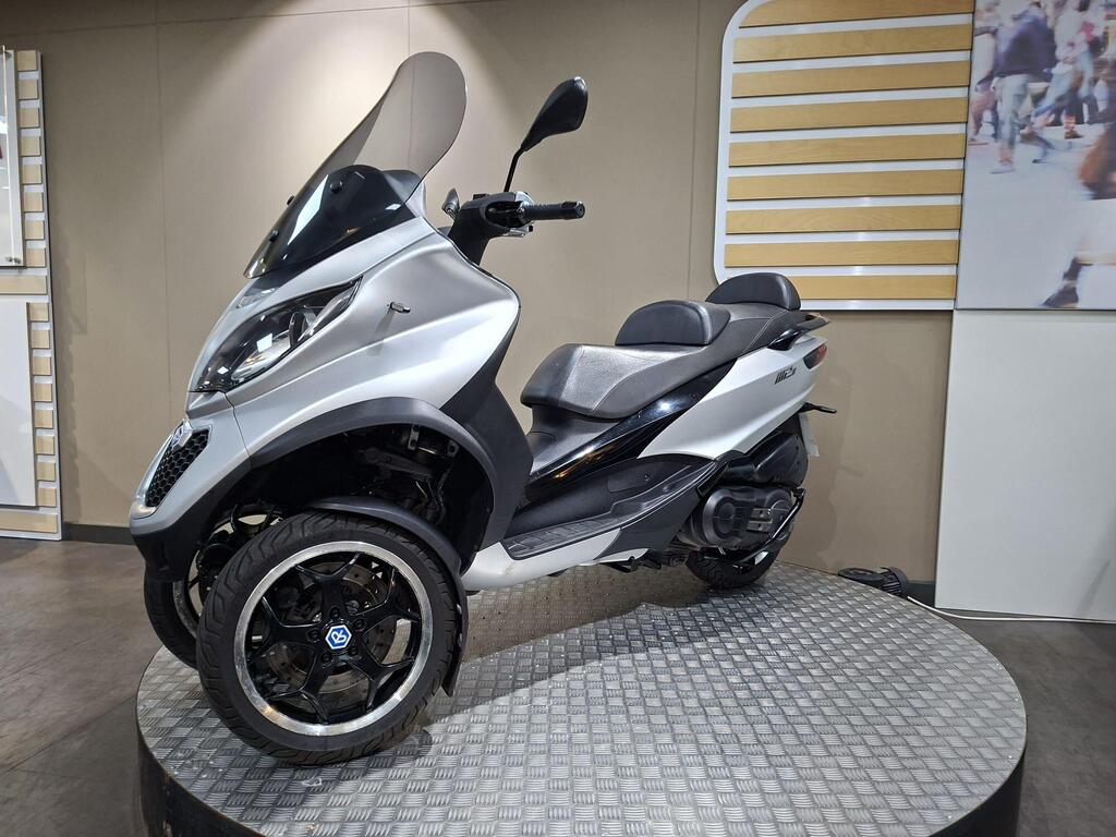 Piaggio MP3 500 2016 17 A silver three-wheeled scooter showcased on a circular display platform, featuring a large windshield and modern design elements. The background includes a plain wall with a striped panel and a partially visible image of people walking.