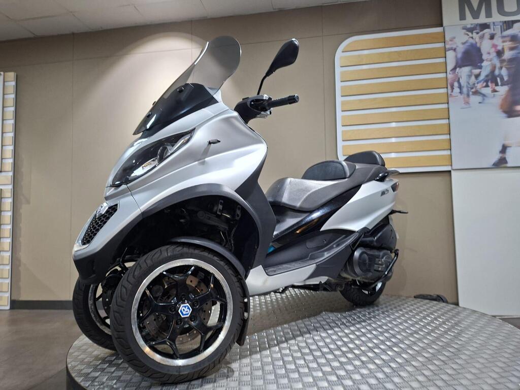 Piaggio MP3 500 2016 18 A silver three-wheeled scooter displayed on a textured platform, featuring a sleek design with a windshield and sporty wheels. The background includes beige walls and a large image of people walking.