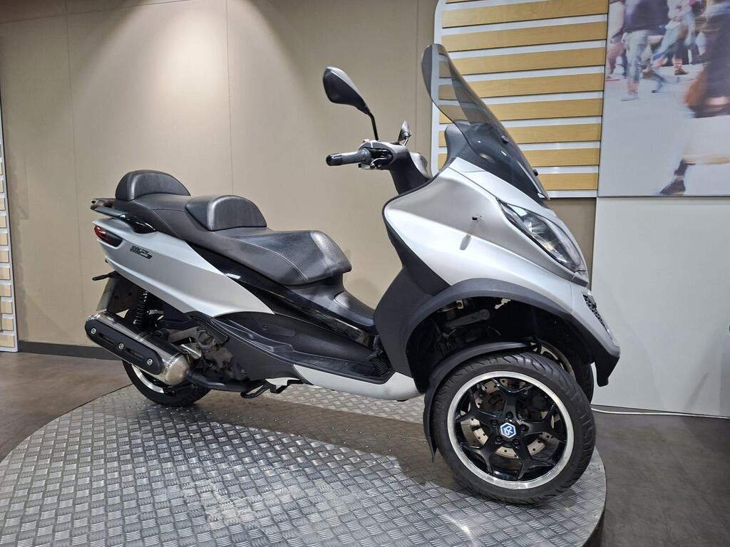 Piaggio MP3 500 2016 2 A modern silver and black scooter with a large windscreen and two seats, displayed on a circular metallic platform against a neutral wall background.
