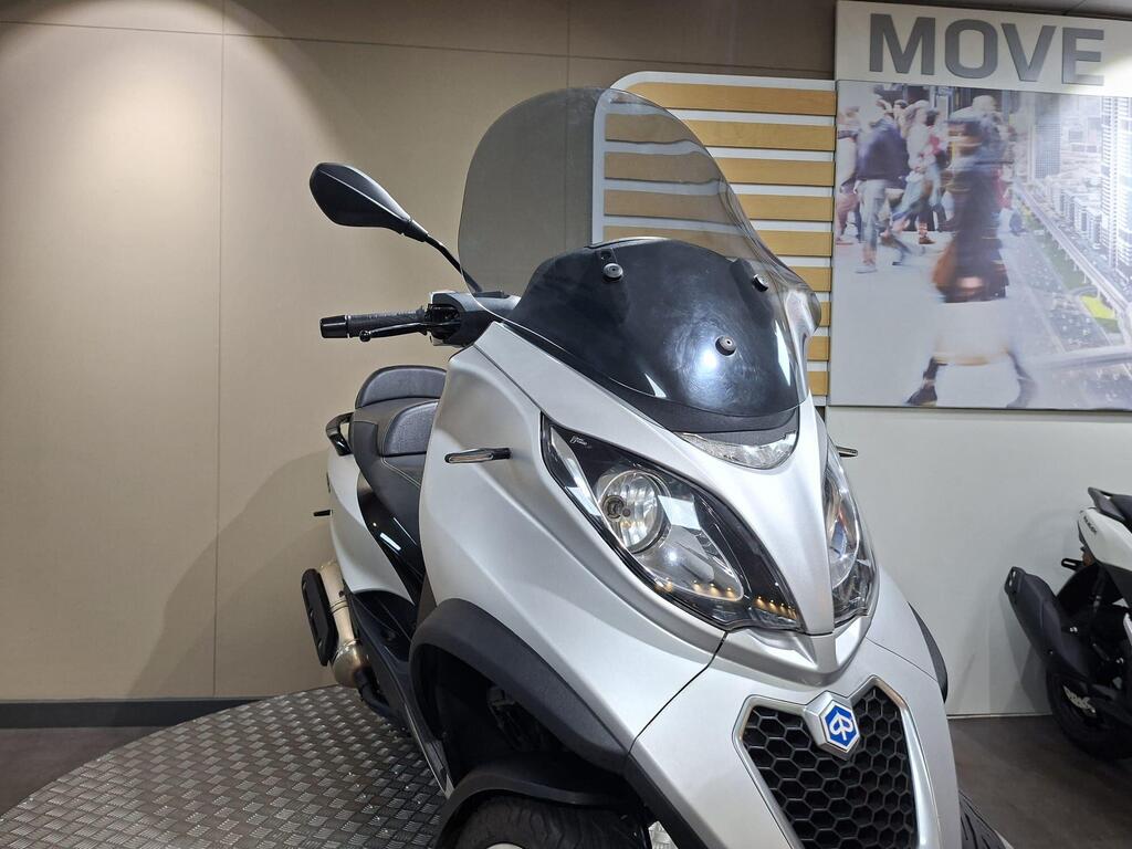 Piaggio MP3 500 2016 20 A silver scooter with a large windshield is displayed in a showroom, featuring a modern design and comfortable seat. In the background, a promotional image depicts people walking in an urban setting.