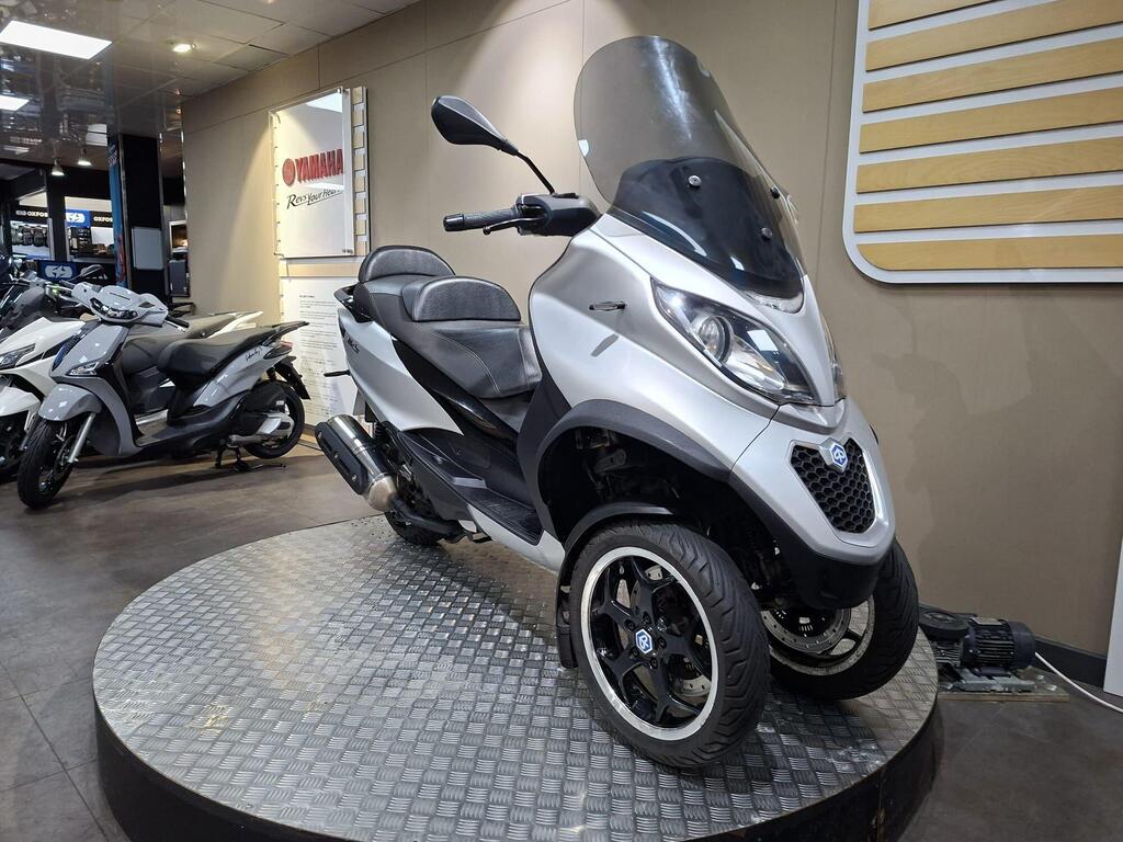 Piaggio MP3 500 2016 4 A sleek silver three-wheeled scooter displayed on a circular platform, featuring a large windshield, comfortable seating, and modern design elements, with additional scooters visible in the background.