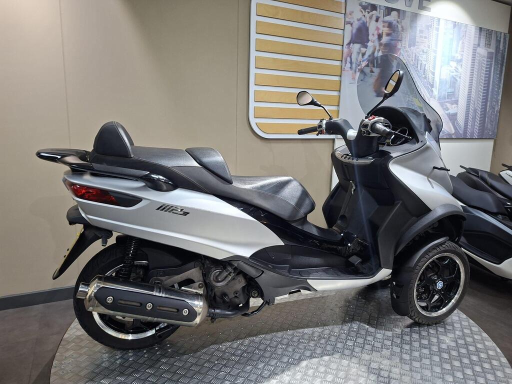 Piaggio MP3 500 2016 6 A sleek silver three-wheeled scooter displayed in a showroom, featuring a black seat, rearview mirrors, and modern design elements, set against a neutral background with a cityscape image.