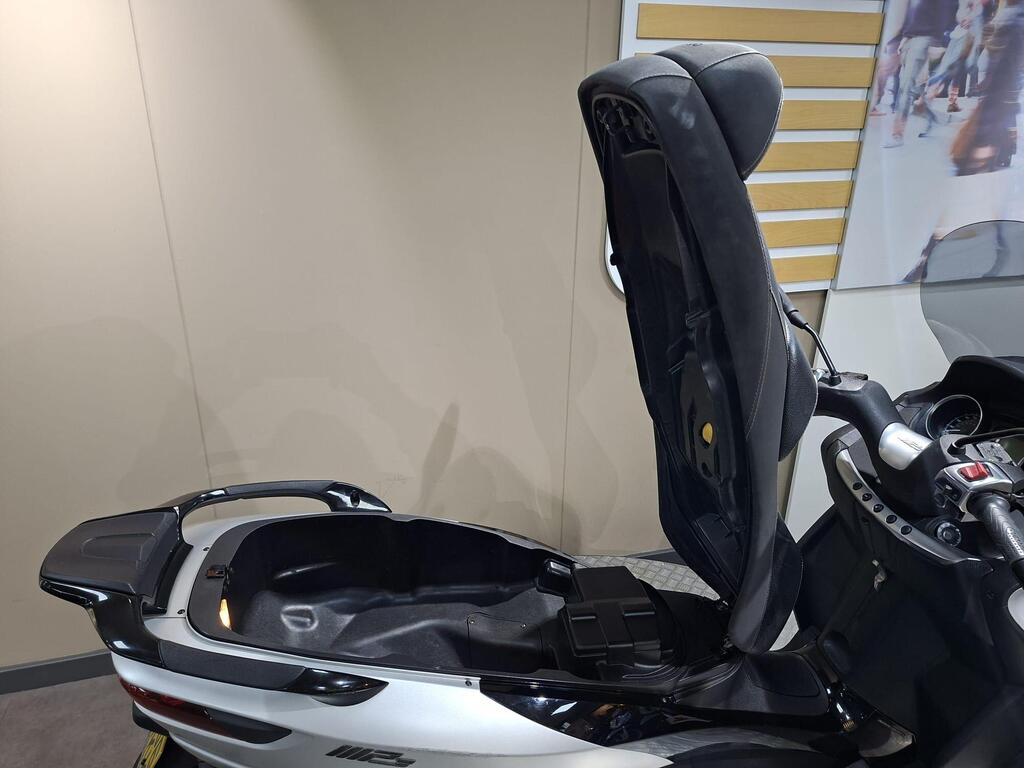 Piaggio MP3 500 2016 8 A side view of a motorcycle with its seat raised, revealing a storage compartment beneath it. The interior of the compartment is black and smooth, set against a neutral-colored background.