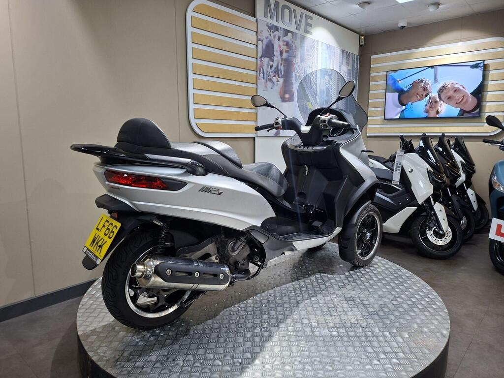 Piaggio MP3 500 2016 9 A silver scooter displayed in a showroom, positioned on a circular platform, with various other scooters in the background and a large screen showing smiling people on the wall.