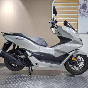 Honda PCX 125 (2024) A sleek gray scooter displayed on a circular platform with a textured surface, showcasing its modern design and prominent features, set against a neutral wall background.