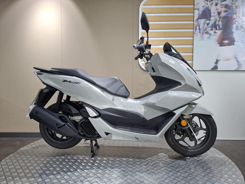 Honda PCX 125 2024 1 Honda PCX 125 (2024) A sleek gray scooter displayed on a circular platform with a textured surface, showcasing its modern design and prominent features, set against a neutral wall background.