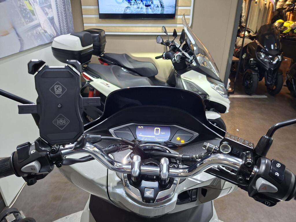 Honda PCX 125 2024 11 A close-up view of a scooter's handlebars and dashboard, showcasing a digital speedometer displaying 0 mph, multifunction controls, and a phone holder. In the background, several scooters and motorcycles are visible.