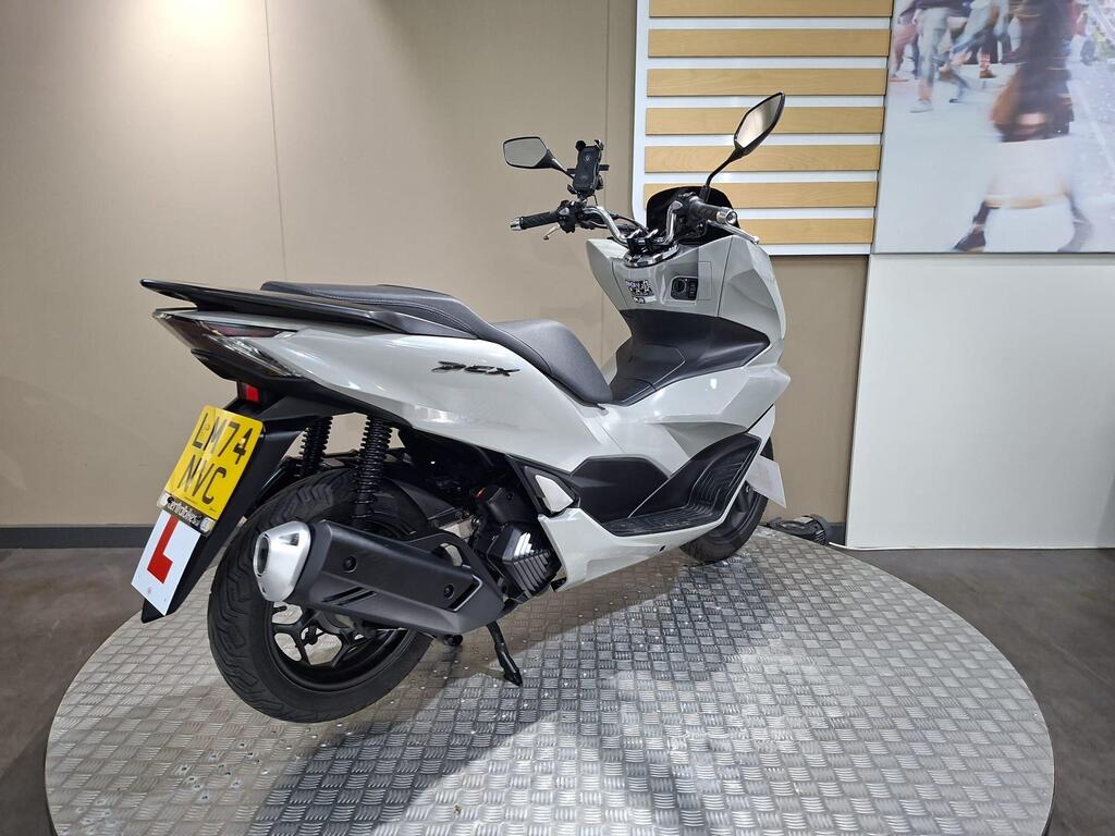 Honda PCX 125 2024 12 A silver motor scooter displayed on a circular platform with diamond-patterned flooring, featuring a sleek design and a license plate indicating it is a learner vehicle. In the background, a wall displays an image of people walking.