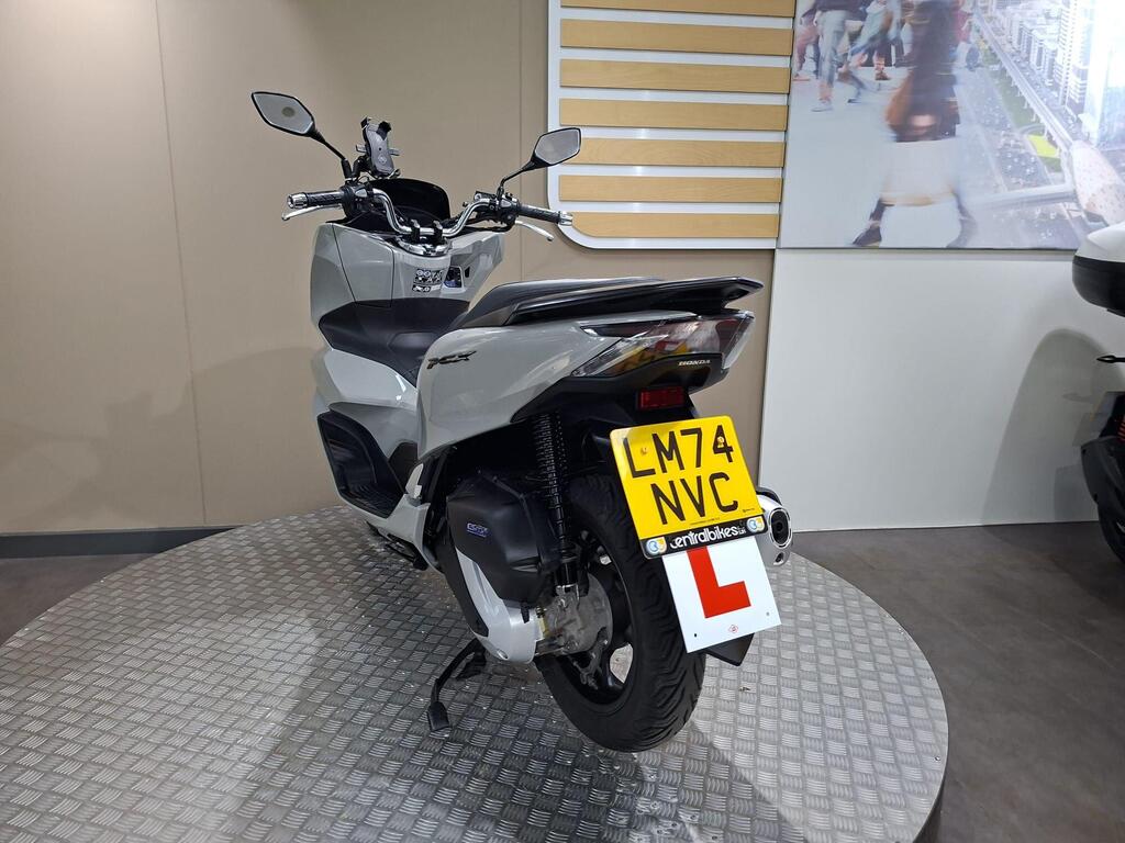 Honda PCX 125 2024 14 A gray Honda motorcycle displayed on a circular platform, featuring a learner's plate with the registration number LM74 NVC. The background includes a beige wall and a large image of a city scene.