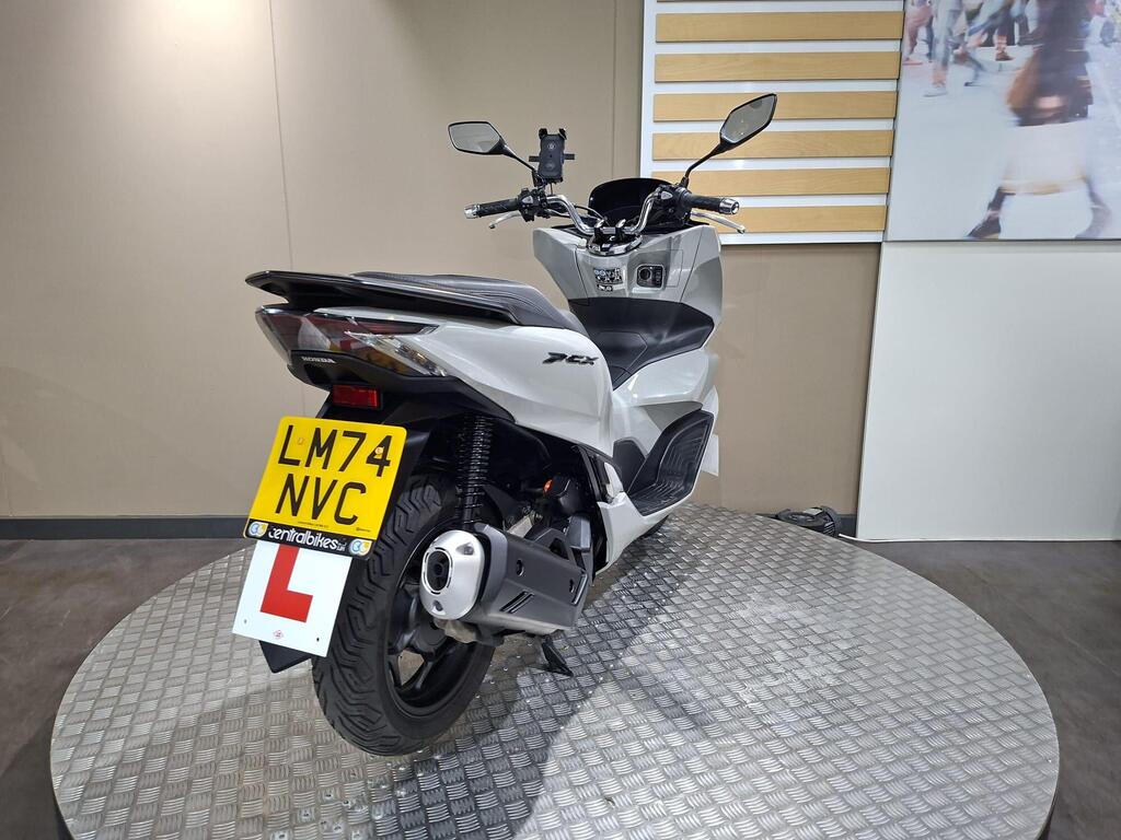 Honda PCX 125 2024 15 A white Honda PCX motorcycle displayed on a circular metal platform, with a visible license plate reading "LM74 NVC" and a learner driver sign. The background features a plain wall and a larger image of people in motion.