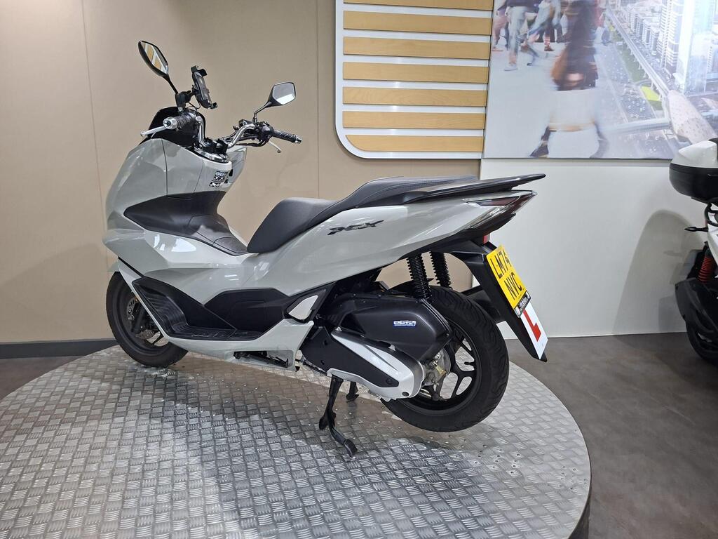Honda PCX 125 2024 16 A sleek, gray scooter displayed on a circular metallic platform in a showroom, with a modern backdrop featuring a cityscape. The scooter is viewed from the rear-left angle, showcasing its streamlined design and distinctive tail light.