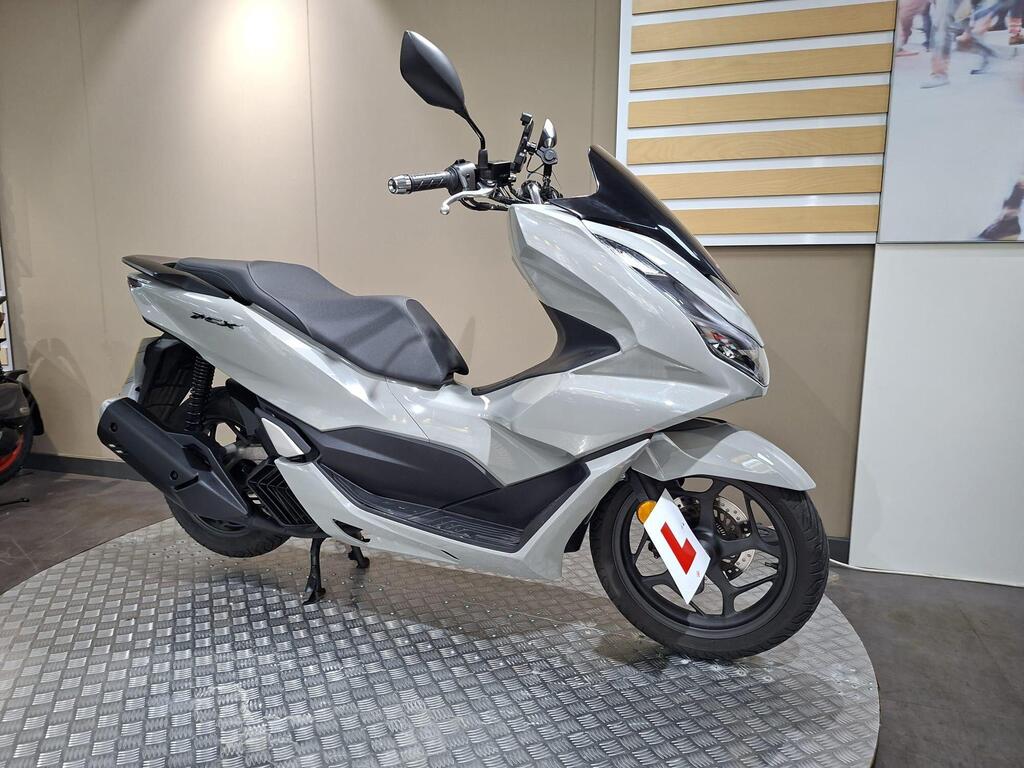 Honda PCX 125 2024 2 A silver scooter with a sleek design is displayed on a circular metal platform in a showroom, featuring a black seat and an "L" learner's permit attached to the side.