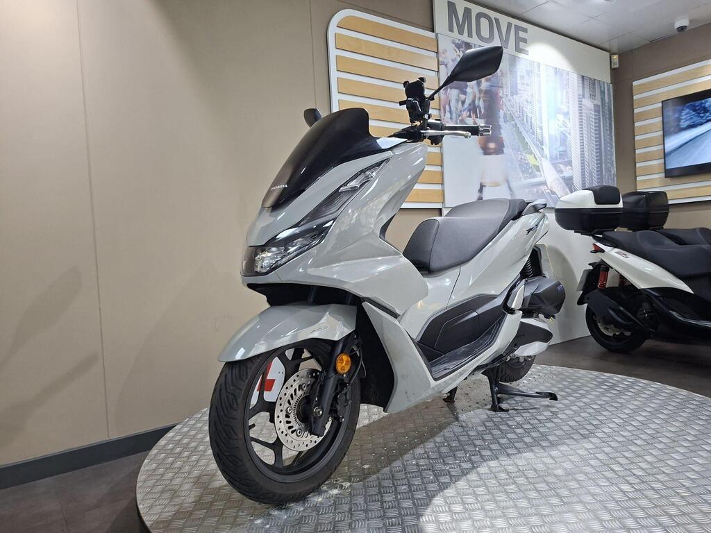 Honda PCX 125 2024 20 A modern gray scooter displayed on a metal platform in a showroom, with a sleek design, visible front wheel, and a digital screen in the background showcasing urban imagery.