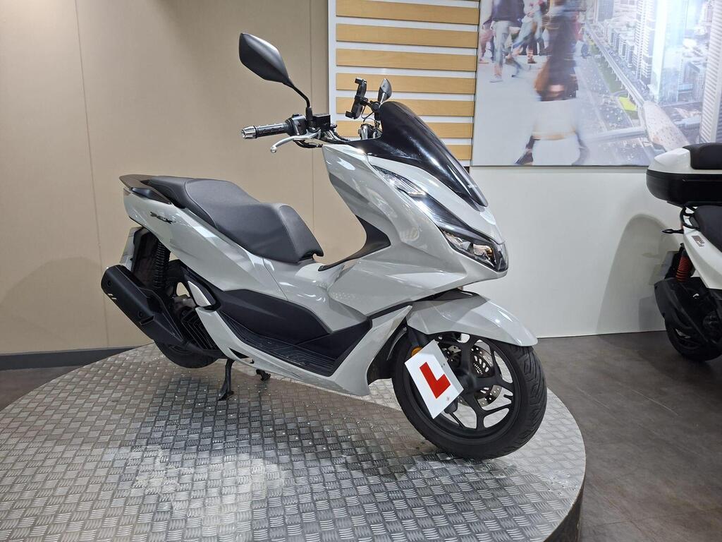 Honda PCX 125 2024 23 A sleek silver scooter with a black seat, featuring an "L" learner's plate, displayed on a circular platform in a showroom with a neutral background.