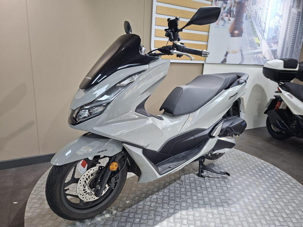 Honda PCX 125 2024 25 A sleek gray scooter displayed on a circular metallic platform, featuring a black windshield and a minimalist design, with a blurred background of an urban setting.