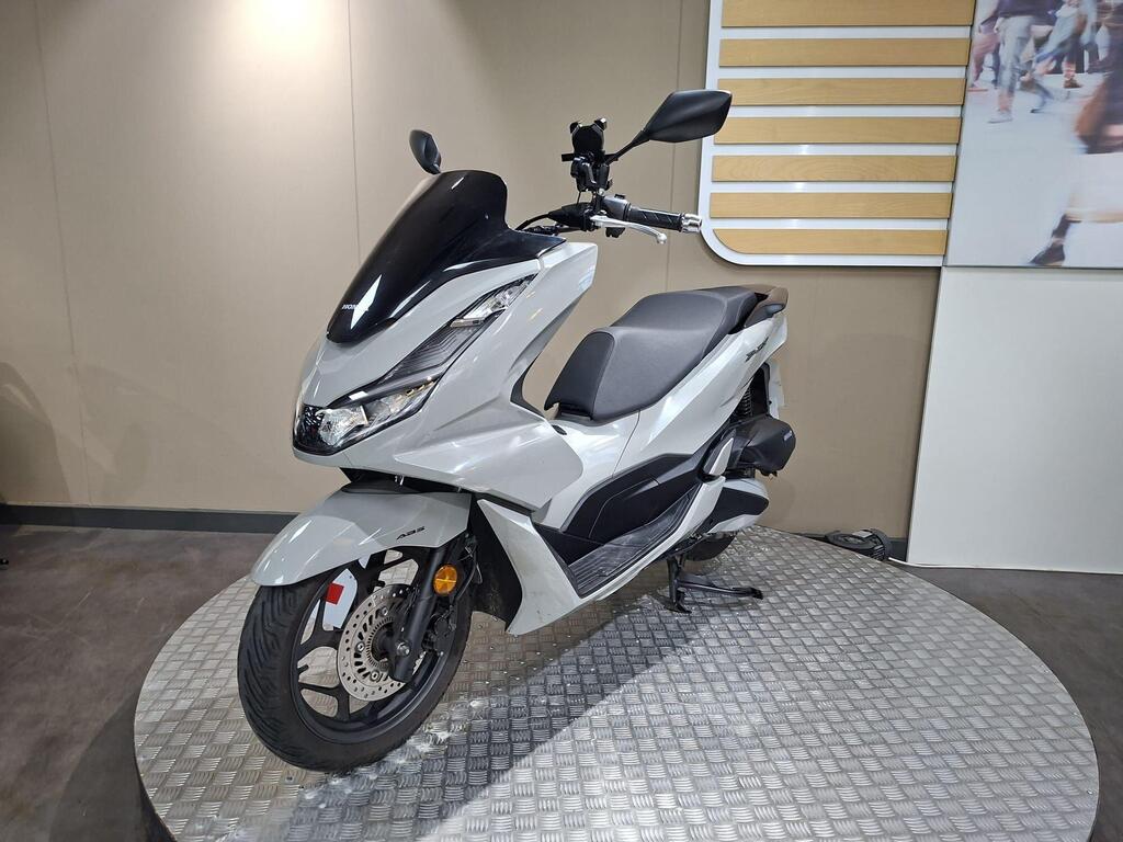 Honda PCX 125 2024 27 A sleek, modern grey scooter displayed on a circular platform, featuring a black windshield, a comfortable seat, and intricate wheel details. The background includes a neutral-colored wall with a partially visible image of people in motion.
