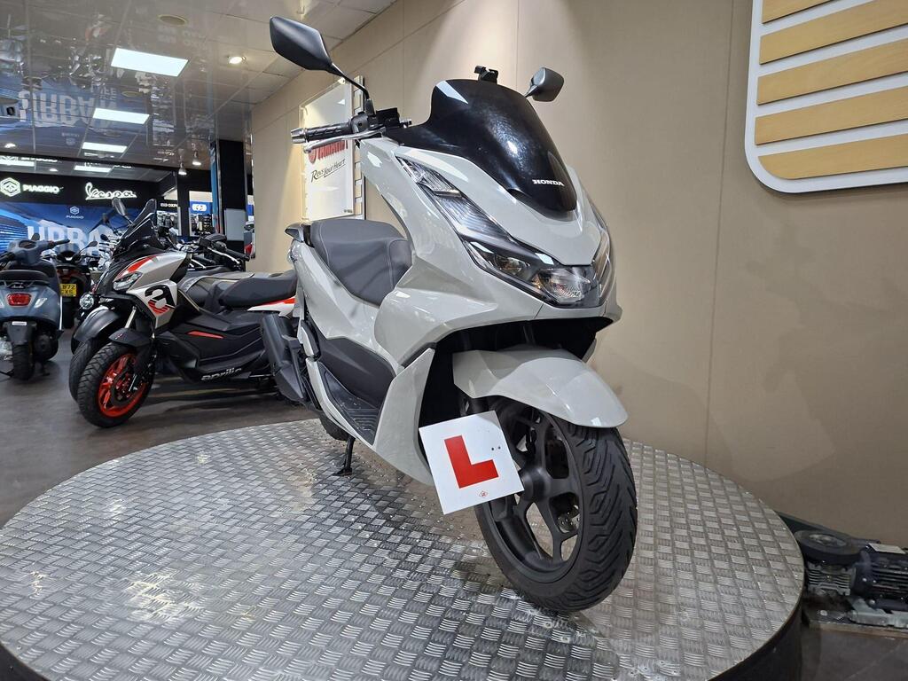 Honda PCX 125 2024 3 A light grey Honda scooter with a learner's plate stands on a circular display in a motorcycle showroom, surrounded by other motorcycles.
