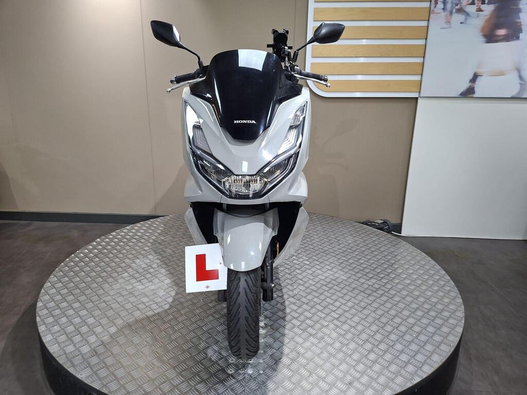 Honda PCX 125 2024 31 A front view of a silver Honda scooter displayed on a circular platform, featuring a learner plate. The background is neutral with a light-colored wall and a partial view of a screen showing people.