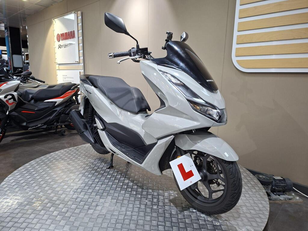 Honda PCX 125 2024 4 A sleek, modern white scooter with a prominent front shield, displayed on a checkered platform in a Yamaha showroom, featuring a learner plate on the front.