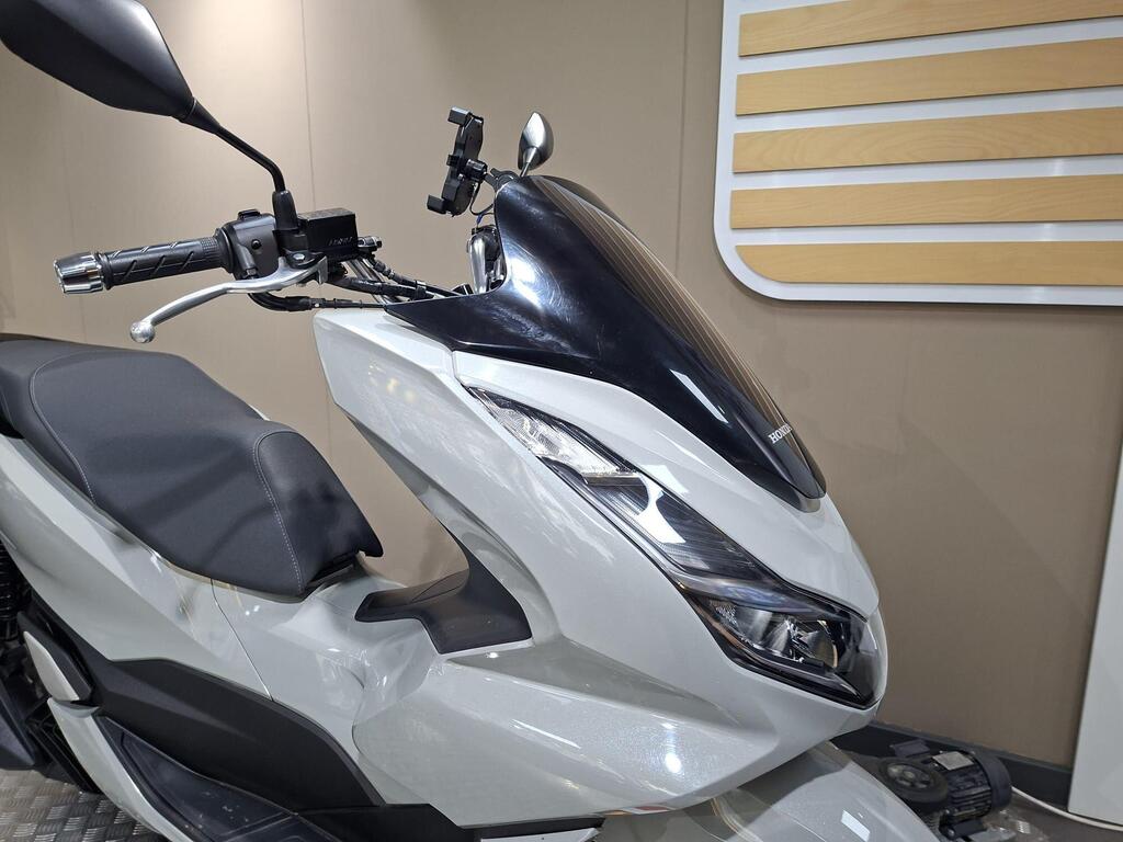 Honda PCX 125 2024 5 A close-up view of a silver Honda scooter, showcasing the handlebars, seat, and aerodynamic front design with a dark windshield. The background features a neutral-toned wall with wooden panel accents.