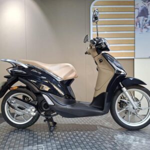 Piaggio Liberty 50 (2020) A sleek black and beige scooter displayed in a showroom, showcasing its modern design and features, with a light-colored wall and an abstract background.