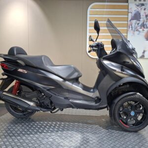 Piaggio MP3 500 Sport (2019) A black three-wheeled scooter with a streamlined design is displayed on a circular metallic platform against a neutral background.