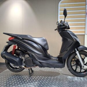Piaggio Medley S 125 (2023) A side view of a sleek black scooter displayed on a textured surface, with a red shock absorber visible and a light-colored wall in the background.