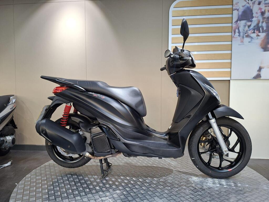 Piaggio Medley (2022) 1 Piaggio Medley S 125 (2023) A side view of a sleek black scooter displayed on a textured surface, with a red shock absorber visible and a light-colored wall in the background.