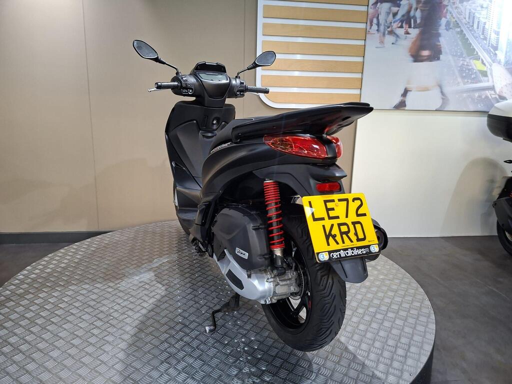 Piaggio Medley (2022) 11 A sleek black scooter displayed from the rear, showcasing its modern design, with a visible license plate reading "LE72 KRD" on a textured circular platform against a neutral background.