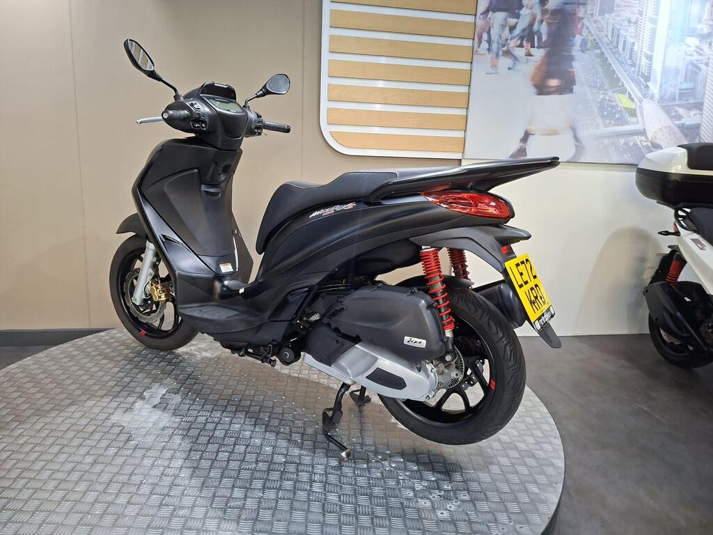 Piaggio Medley (2022) 12 A sleek black scooter displayed on a circular metal platform, showcasing its sporty design and red rear suspension, with a blurred, modern city backdrop.