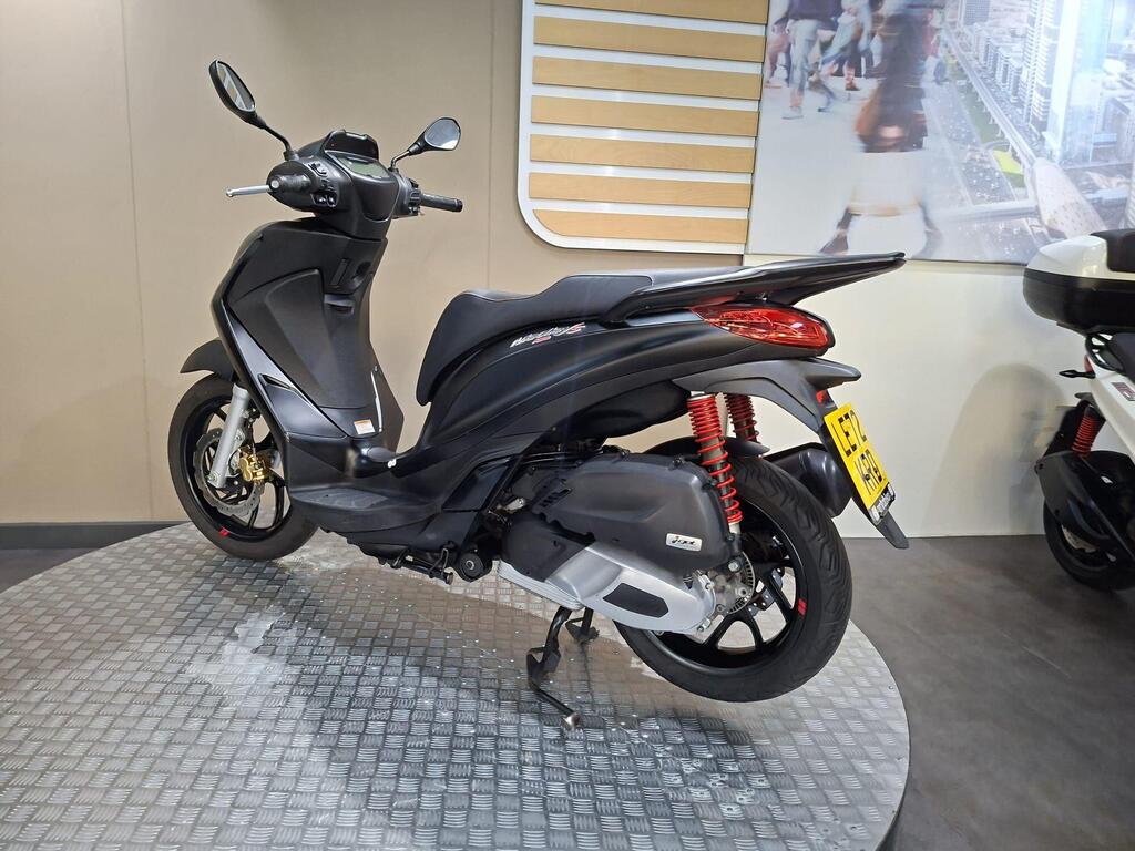 Piaggio Medley (2022) 13 A sleek black scooter is displayed on a circular platform in a modern showroom, with a blurred cityscape visible in the background. The scooter features red suspension springs and a prominent rear light.