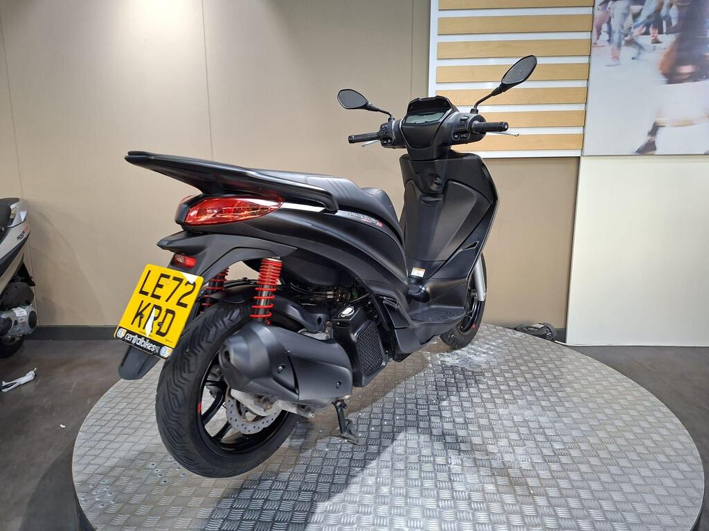 Piaggio Medley (2022) 14 A black scooter with red shock absorbers is displayed on a rotating platform against a neutral background, showcasing its sleek design and features. The license plate reads LE72 KPD.
