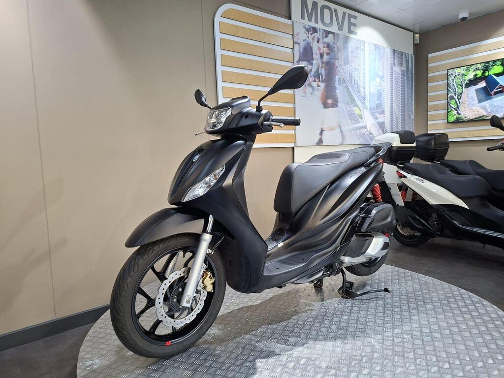 Piaggio Medley (2022) 15 A black scooter displayed on a checkered floor, with a modern interior featuring a wall banner that says "MOVE" and an LCD screen showing green outdoor images in the background.