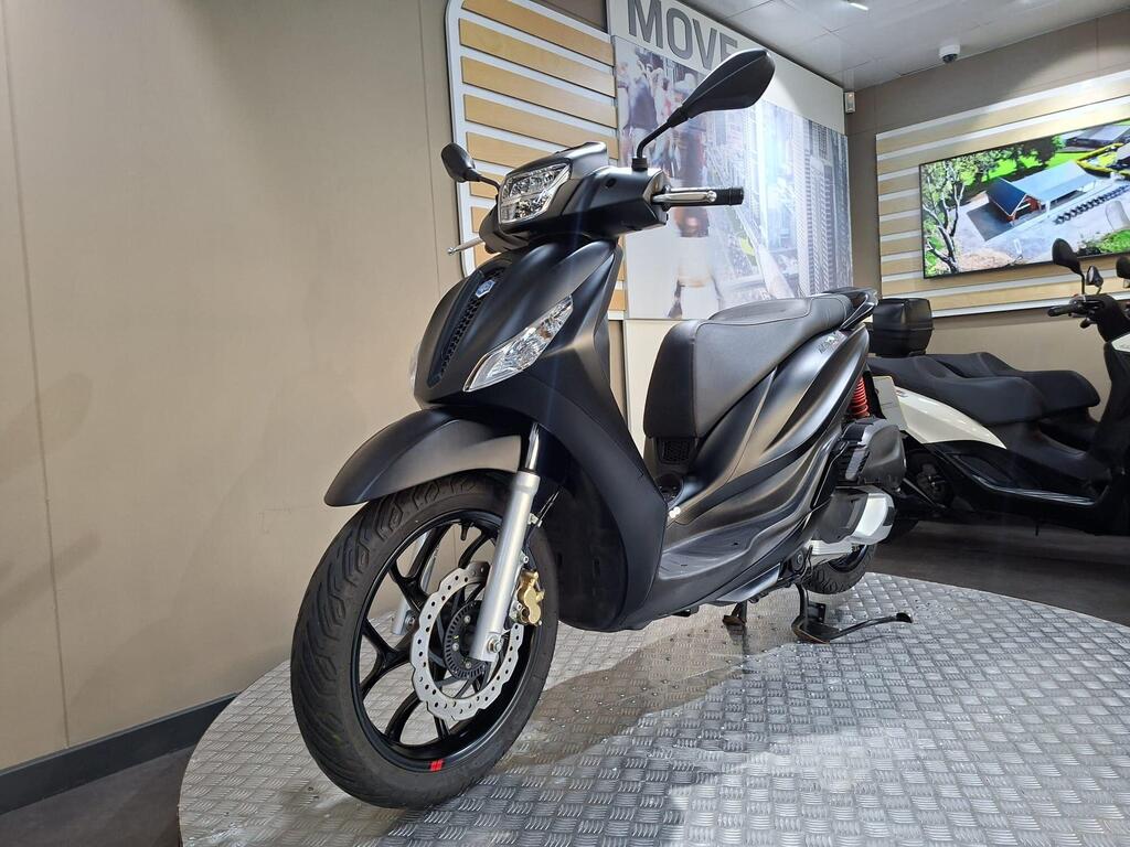 Piaggio Medley (2022) 16 A black scooter displayed on a circular platform in a showroom, with a modern backdrop featuring digital screens and a light-colored wall.